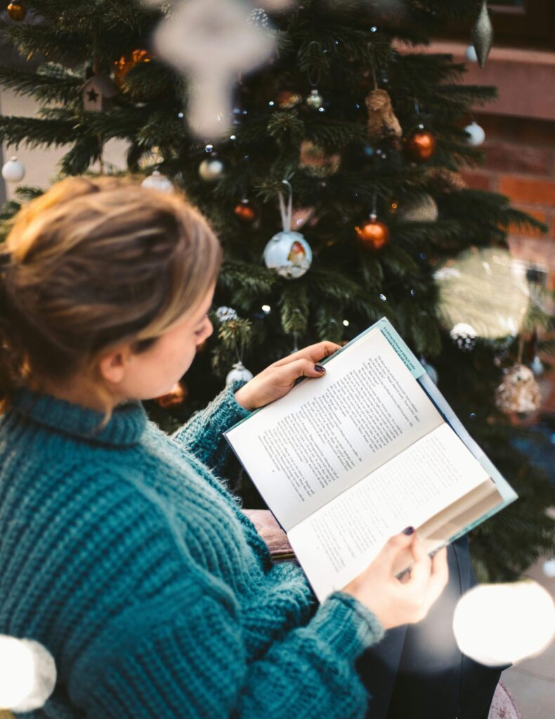 A woman reading near a Christmas tree with a cup of tea and relaxing, practicing self-care during the holidays.